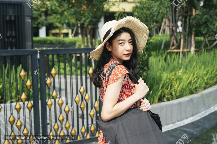 年轻漂亮的泰国亚裔旅行家女子身穿红色连衣裙，头戴红帽，在曼谷市公园旅游。