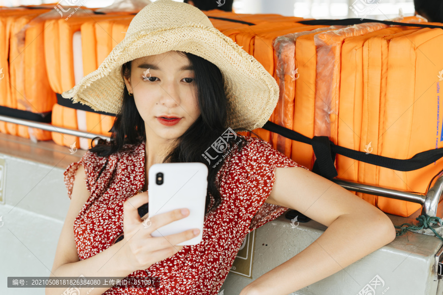 年轻漂亮的泰国亚裔旅行家，穿着红裙子，戴着红帽子，坐在船上。用智能手机和朋友聊天。