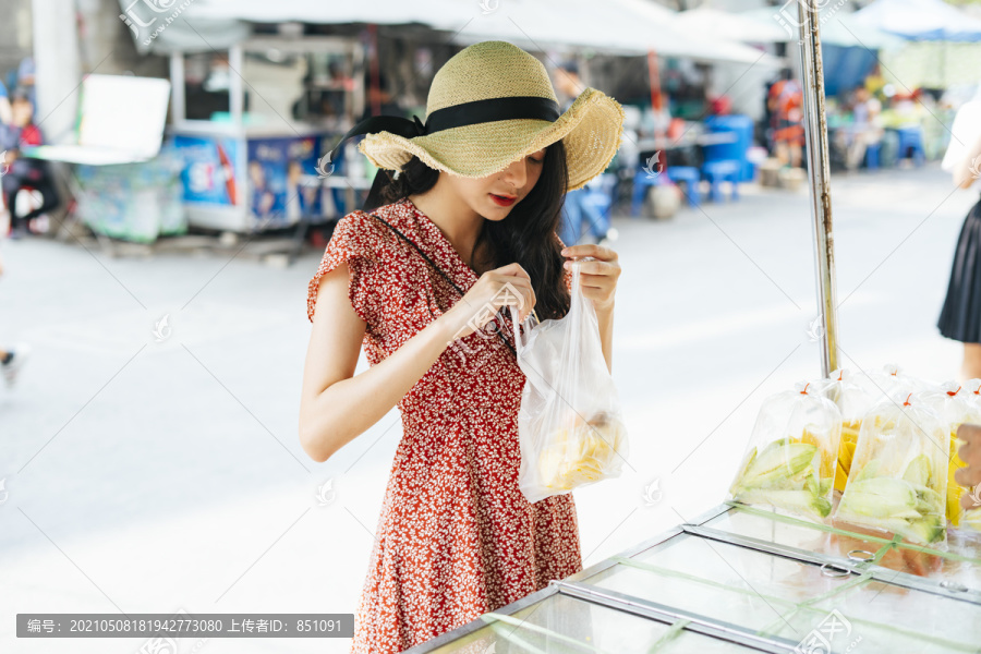 年轻漂亮的泰国亚裔旅行家，穿着红裙子，戴着红帽子，从街边小摊上买水果。