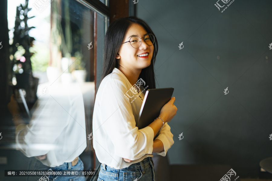 开朗美丽的亚洲女人戴着眼镜，抱着笔记本电脑站在镜子窗前。