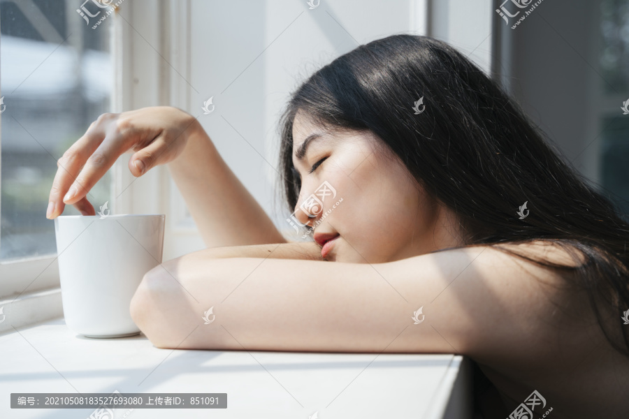 长发女孩坐在窗前，在晨光下端着一杯咖啡。