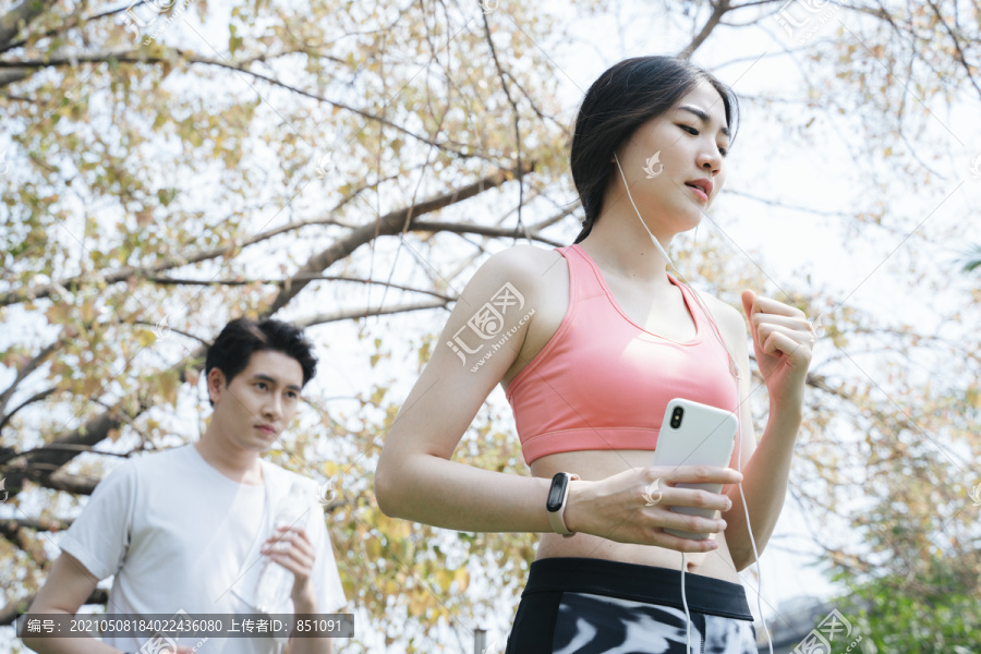 一个长发女人穿着旧的玫瑰色运动胸罩在秋天的公园里跑步，一个穿着白色t恤的男人在她身后跑。