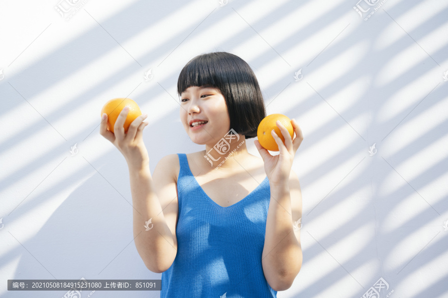 开朗快乐美丽的鲍勃发型穿着蓝色背心衬衫的亚洲女人手拿两个橘子热带水果在白色背景上。