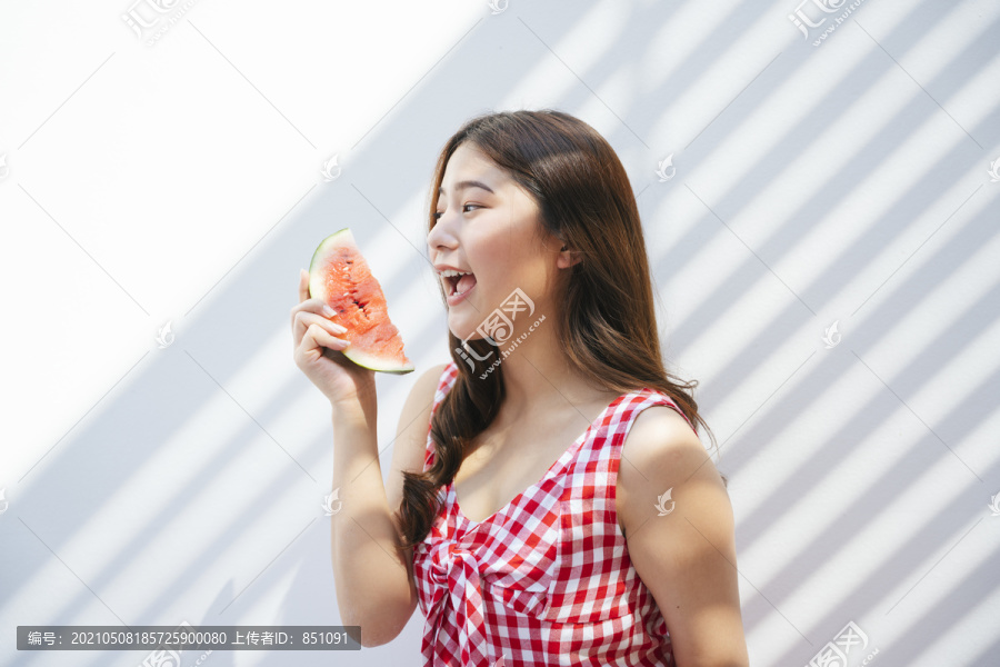 一位身穿红色连衣裙的亚洲年轻女子在背景上吃着新鲜的西瓜。带着水果的快乐女人。