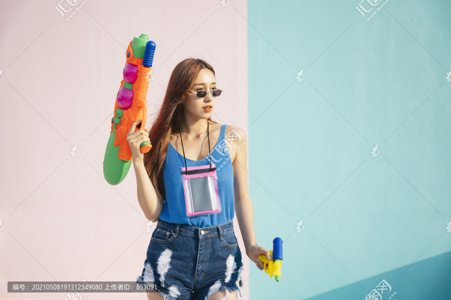 身着蓝色背心衬衫、戴墨镜、手持两支水枪的性感女子肖像。宋克兰节的概念。在柔和的蓝粉色背景上孤立。曼谷