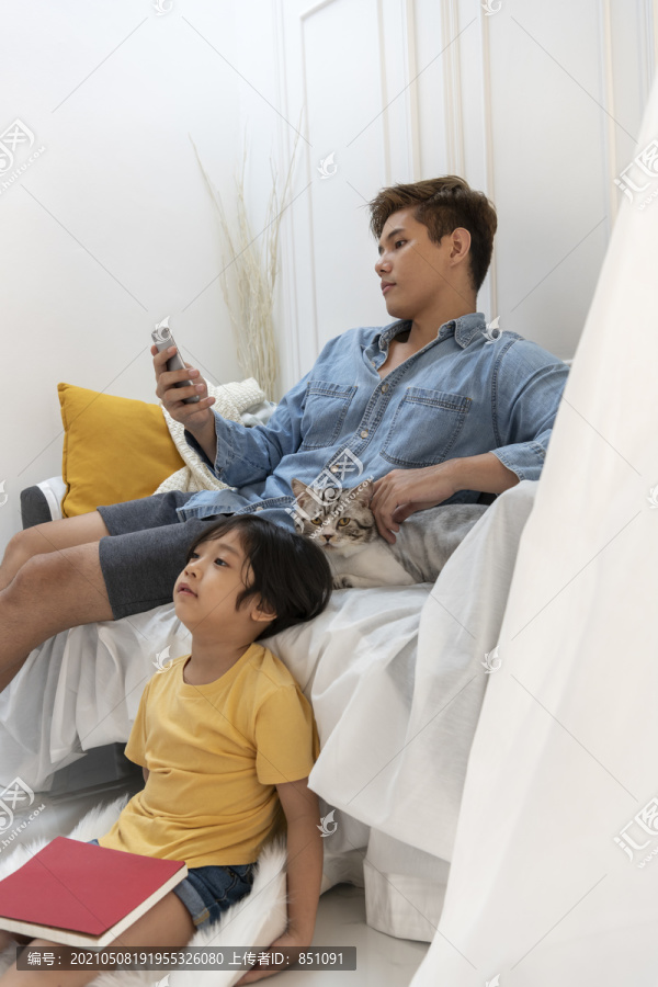 快乐的亚洲家庭和猫住在一起。爸爸用智能手机和猫摩擦。儿子厌倦了看书，看电视。