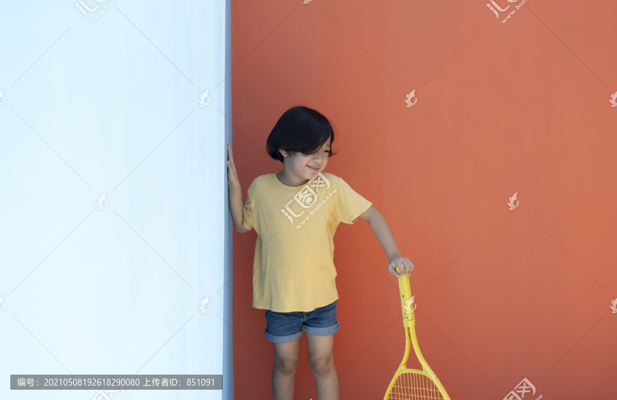 穿着黄色衬衫的可爱小男孩拿着网球拍，背景是五颜六色的蓝红色。害羞的男孩拿着网球拍。