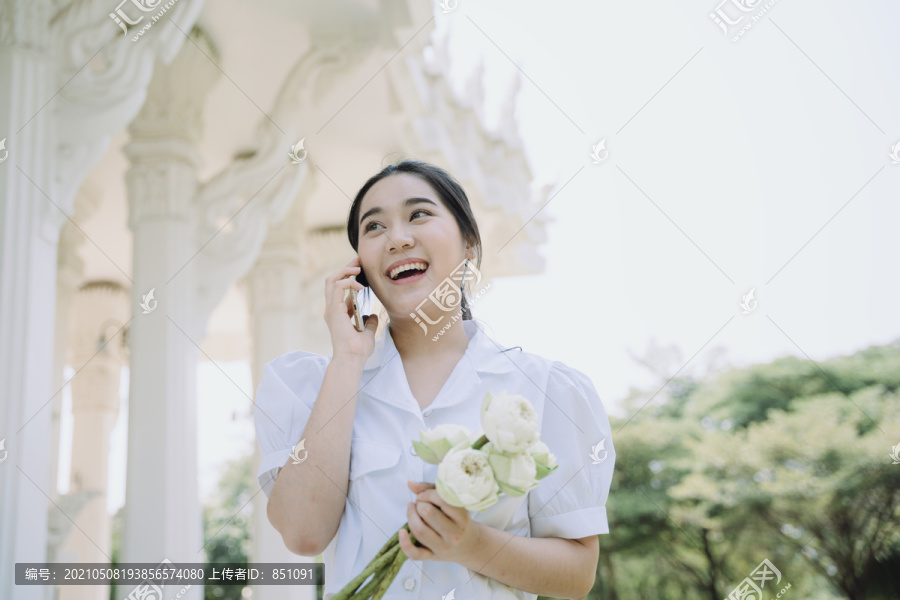 穿白衬衫的黑马尾女孩一边接电话，一边抱着荷花走向寺庙。