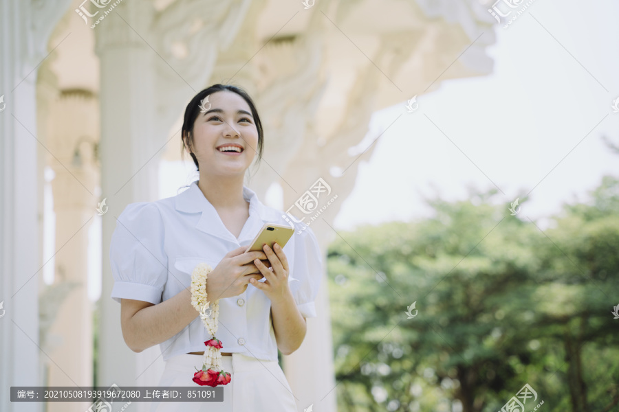 身穿白衬衫的黑马尾海特女孩把茉莉花环挂在手腕上，走在庙里，用智能手机复制空间。