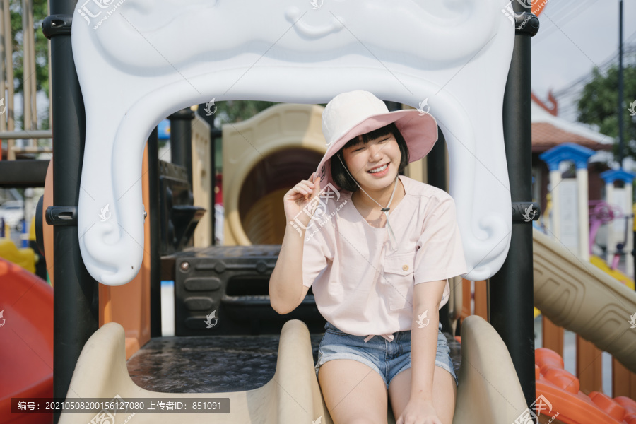 美丽的泰国亚裔女子戴着帽子，暑假喜欢在游乐场公园玩滑梯。