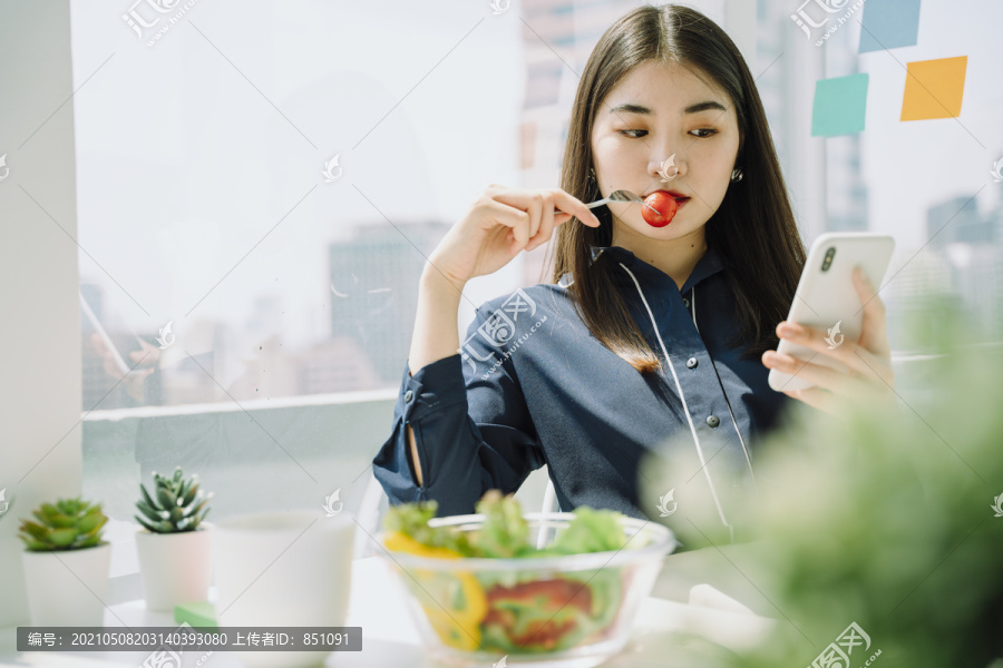 美丽的亚洲年轻泰国女子，深色长发，喜欢在办公室吃沙拉碗和使用智能手机。女商人喜欢蔬菜沙拉碗。午休时间