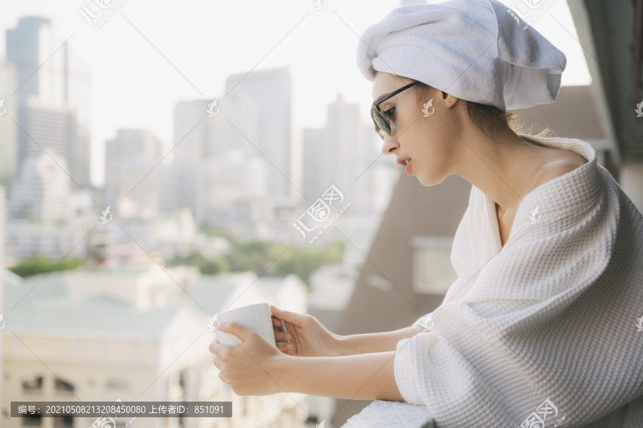 复制空间。侧视图-在公寓阳台，穿着白色浴袍的年轻女子拿着一杯咖啡。