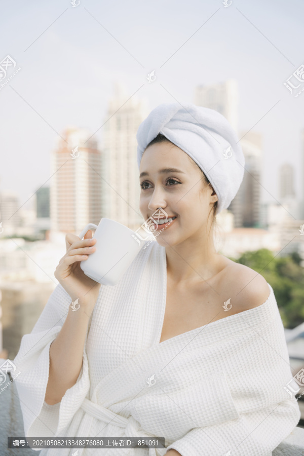 身着白色浴袍的年轻女子在城市背景的公寓阳台上享用热咖啡。