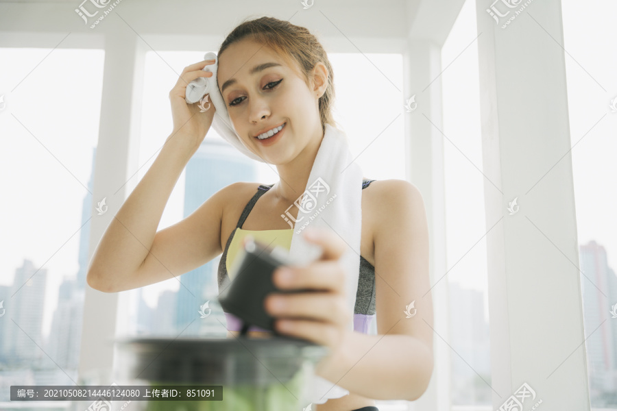 穿着运动内衣和毛巾的美丽健康的女人在家锻炼后，在厨房用搅拌机调制健康的蔬菜汁。
