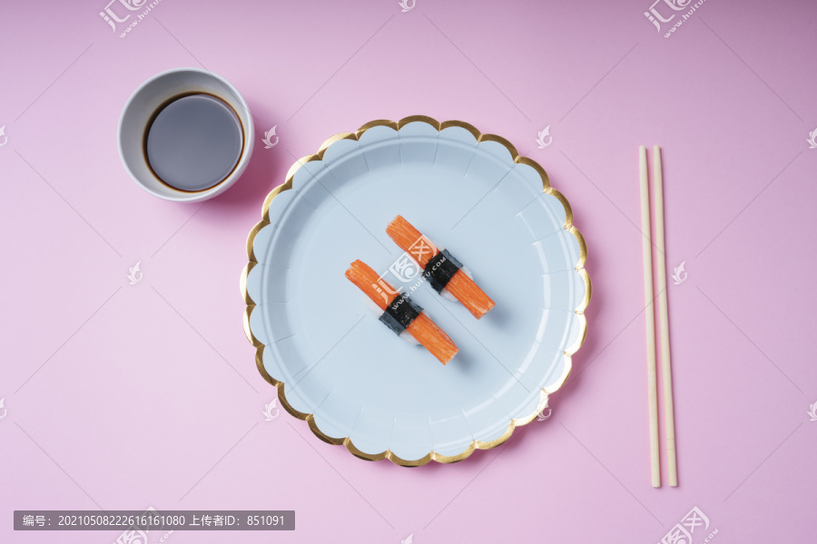 两个蟹棒寿司放在一个盘子里，酱油和筷子放在粉红色的背景上。