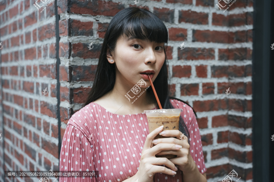 美丽的亚泰长黑发女子喝冰咖啡用吸管在街上砖背景。