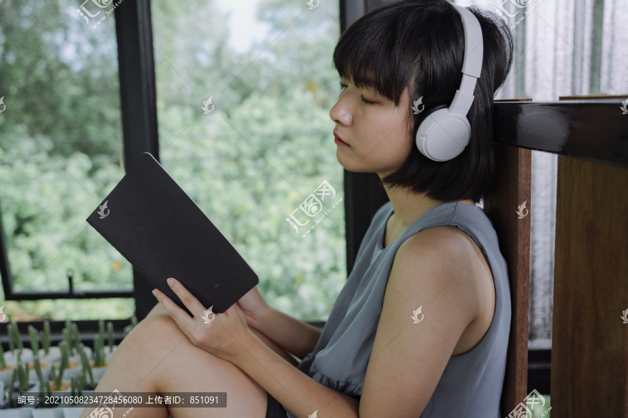 侧视图-美丽的亚泰女人深色短发，戴着耳机听音乐，在当地的旅馆房间看书。