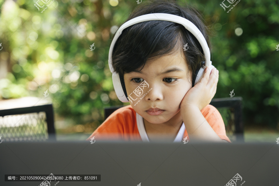 严肃的亚洲小孩在户外院子里戴着耳机用笔记本电脑。