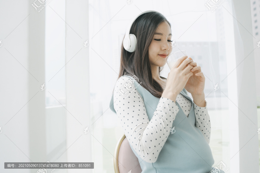 年轻漂亮的亚洲孕妇戴着无线耳机端着一杯牛奶。