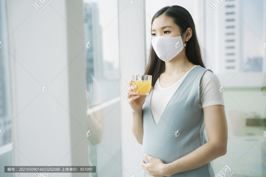 美丽的亚洲孕妇戴着棉质口罩，在家里捧着一杯橙汁。健康饮料。