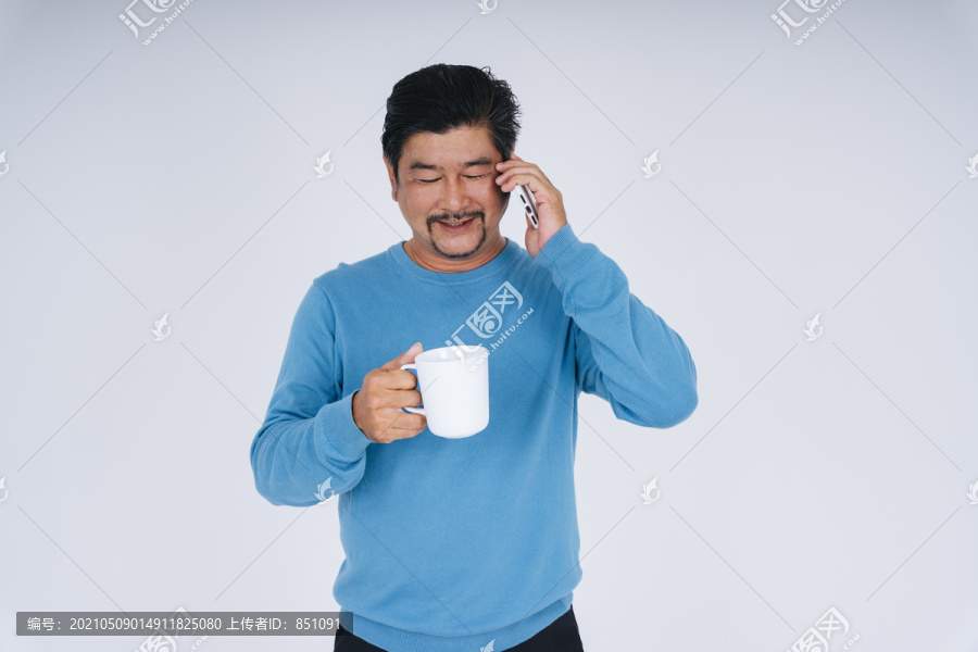 身着蓝色长袖衬衫的老人手持白色咖啡杯在打电话。