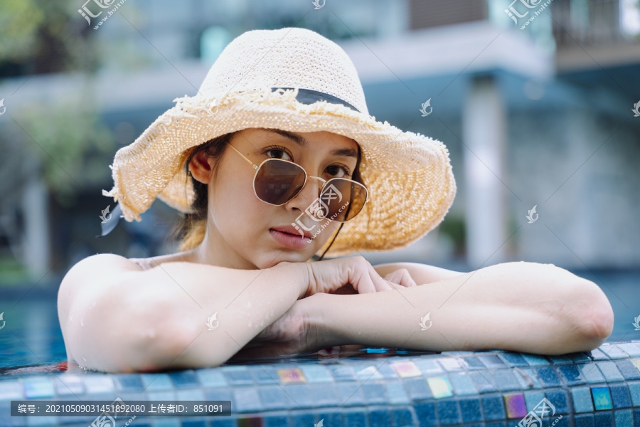 美丽的亚洲女游客戴着墨镜和帽子在游泳池里放松。