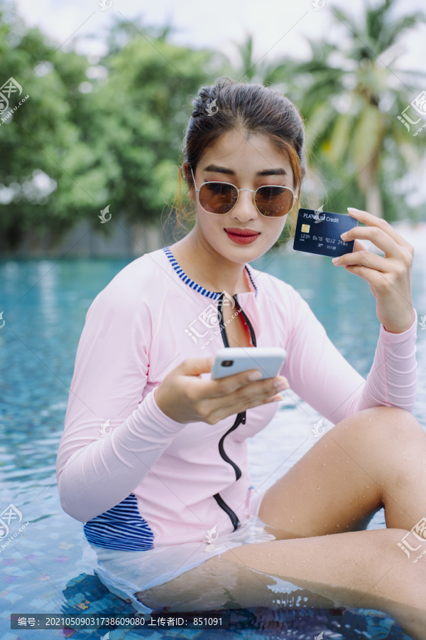 穿着泳衣的亚洲美女游客在室外游泳池持信用卡。