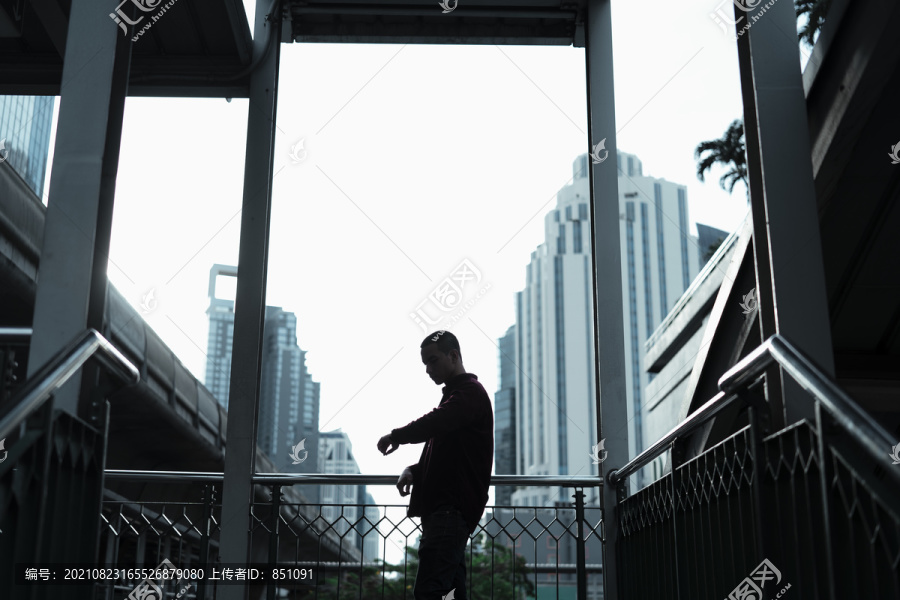 穿着红衬衫的光头党男子站在天桥上，戴着镜框，看着手表，剪影。