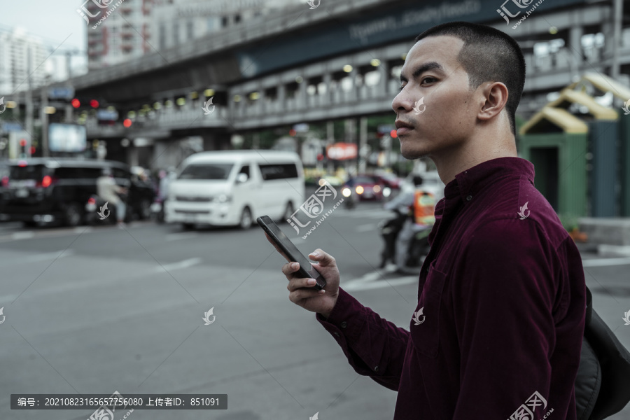 身穿红衫的光头男子在路边手持智能手机，等待穿过马路。