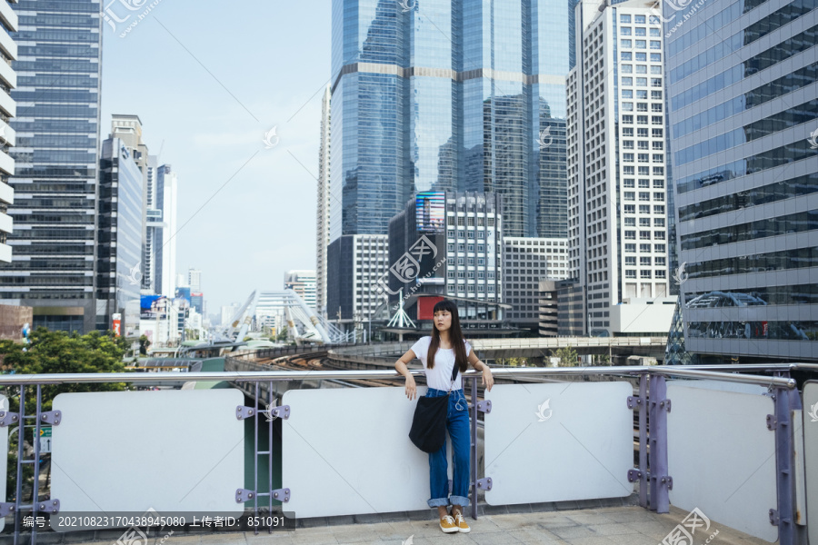 一个留着刘海发型的可爱泰国女人靠在扶手上，等着有人来接她。城市景观背景。