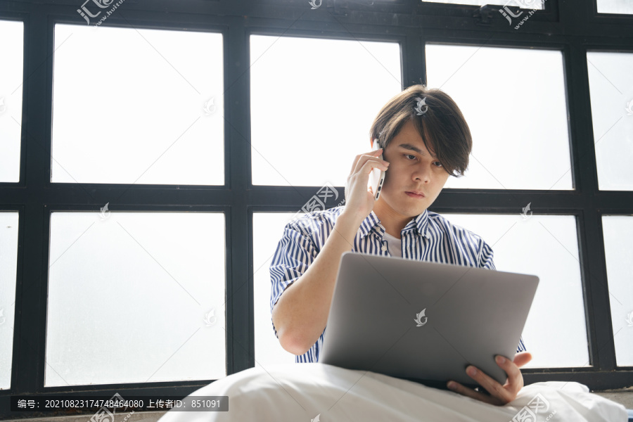 在家里工作的亚洲年轻人，在窗户附近拿着笔记本电脑和智能手机。