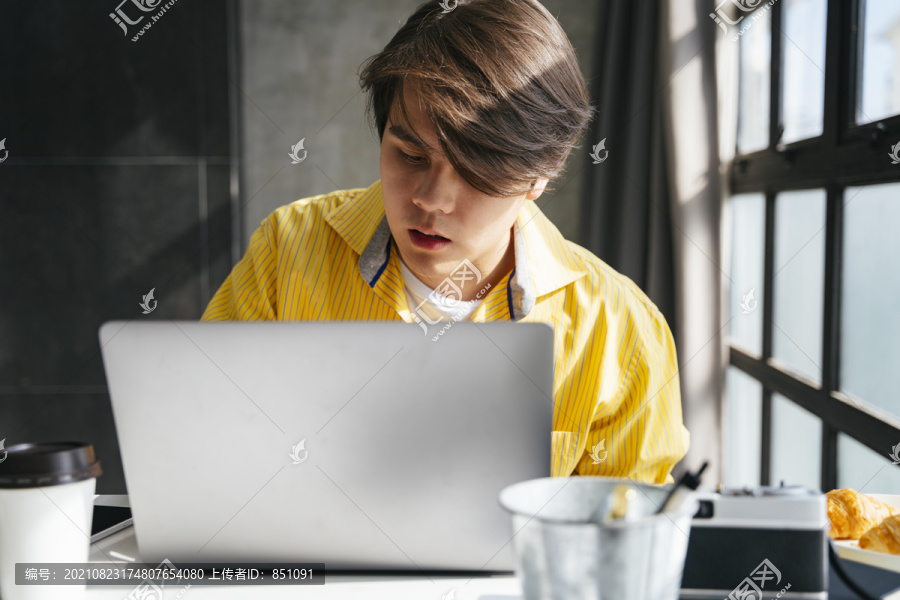 穿着黄衬衫的亚洲年轻人在现代办公室里用笔记本电脑工作。