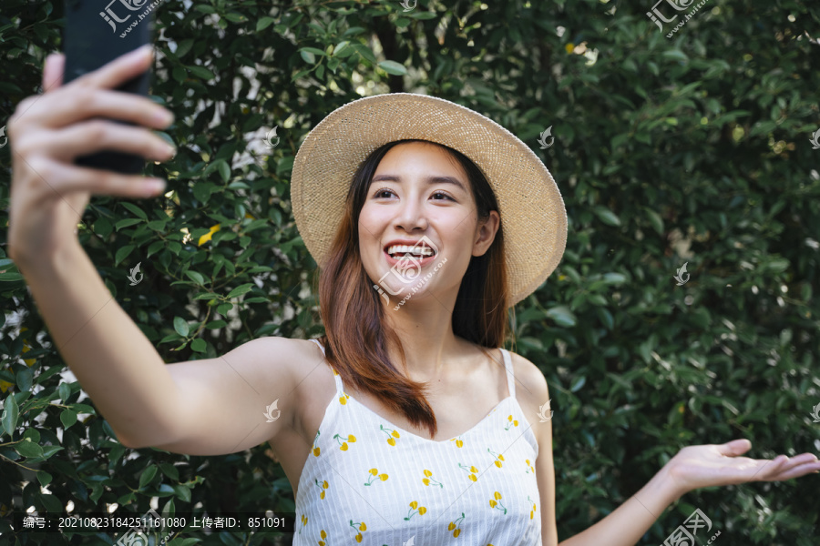 美丽的亚洲女人戴着帽子，穿着白色的裙子，在公园里自拍或者用智能手机进行旅行。