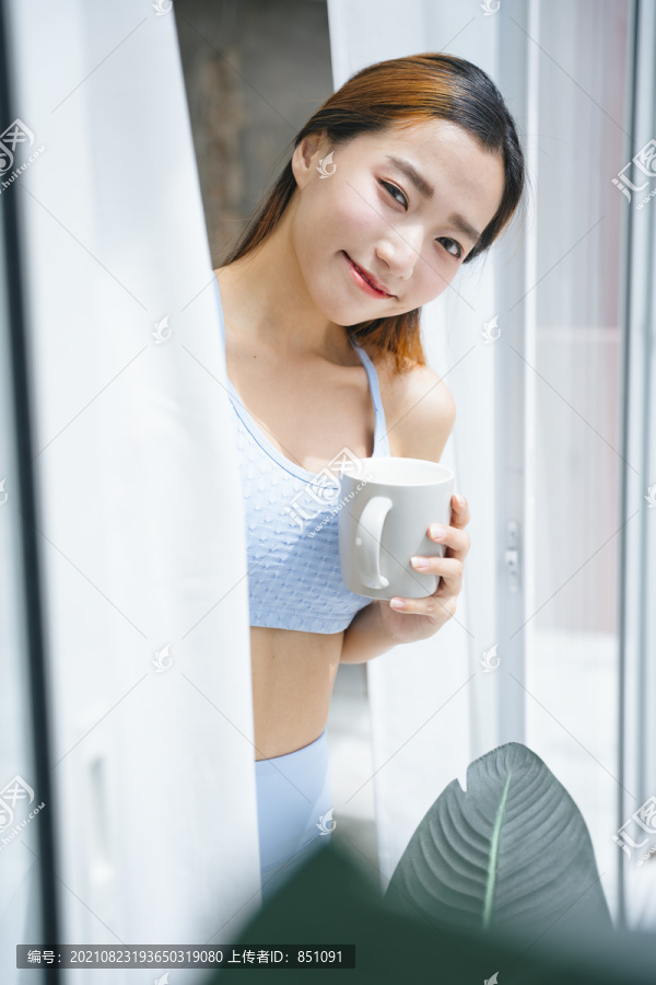 美丽的运动型亚洲女性，穿着运动胸罩，在家阳台上端着一杯咖啡。
