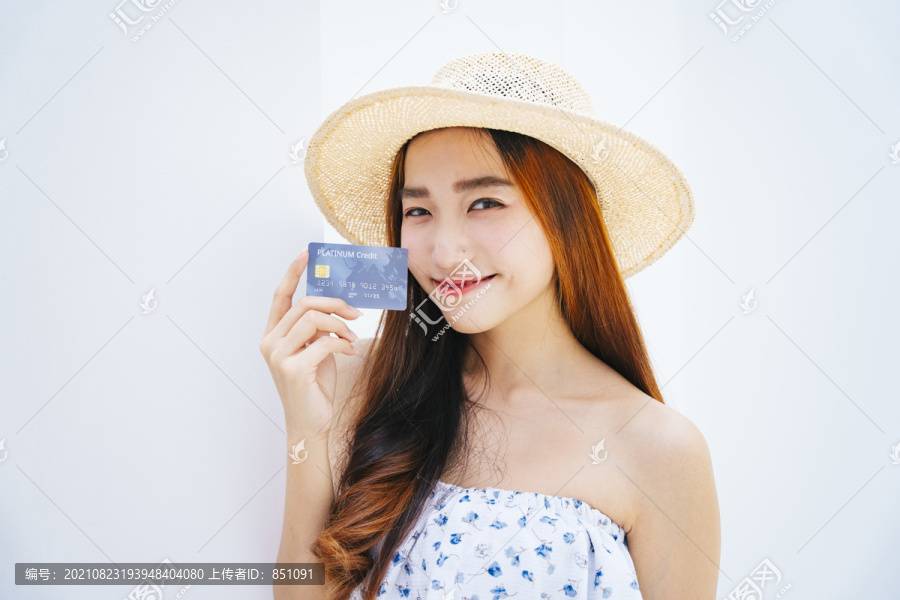 戴着帽子、穿着白色连衣裙、手持信用卡的亚洲美丽年轻女子的肖像。