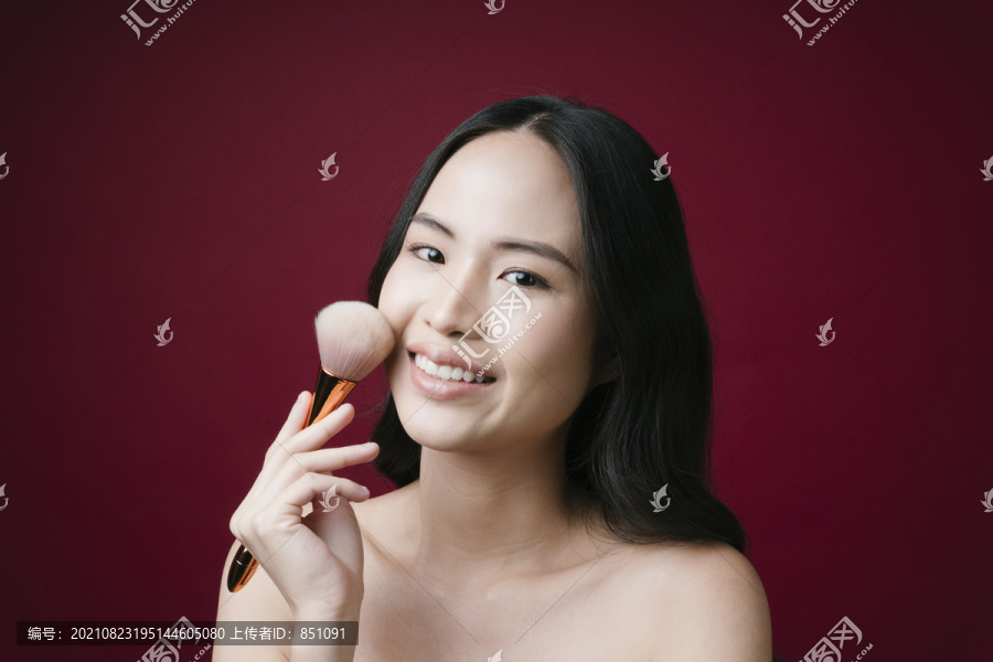 美丽的年轻亚洲女孩的画像，用化妆刷自然化妆，隔离在红色背景上。