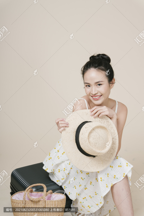 亚洲女旅行者的肖像，拿着草帽坐在行李上。