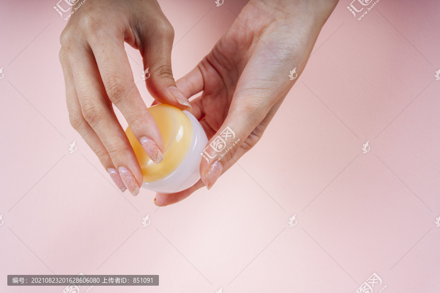 手持粉红色背景上隔离的小罐子面霜乳液的女人的手。