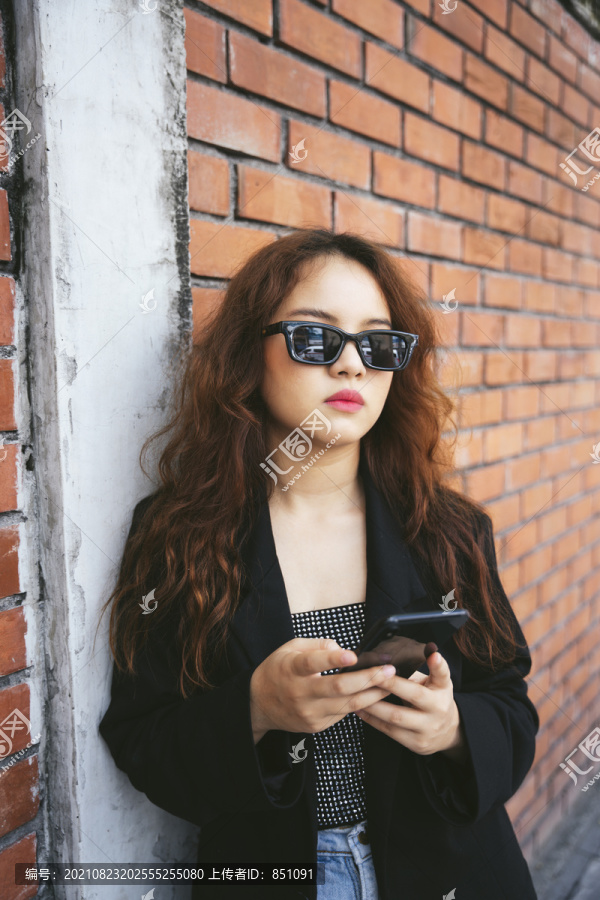 穿着墨镜和黑色夹克的年轻亚洲女性在街上使用智能手机的时尚肖像。