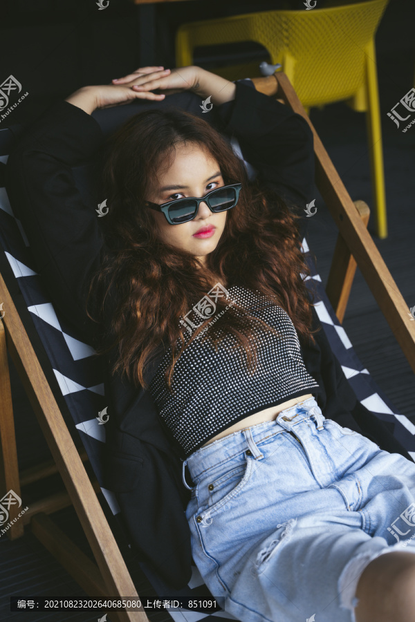 穿着墨镜和黑色夹克坐在沙滩椅上的年轻亚洲女性的时尚肖像。