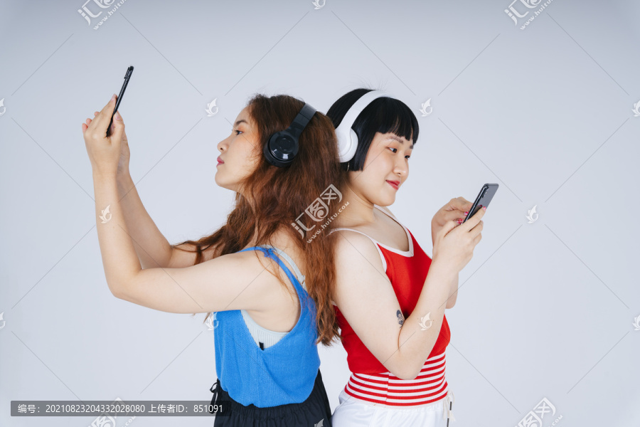两位年轻亚洲女性在白色背景下听音乐和玩手机游戏的肖像。