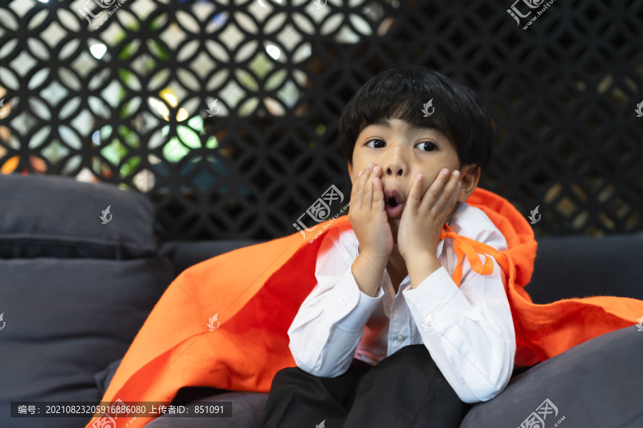 在万圣节派对上，亚洲男孩坐在沙发上，戴着橙色的风帽，打扮得漂漂亮亮。