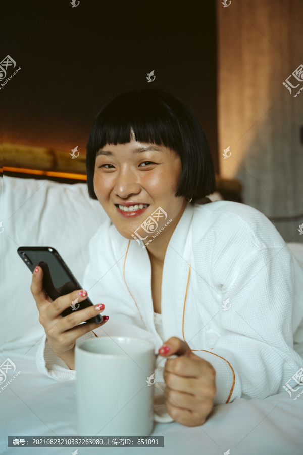 亚洲短发女性在床上使用智能手机，手持咖啡杯。