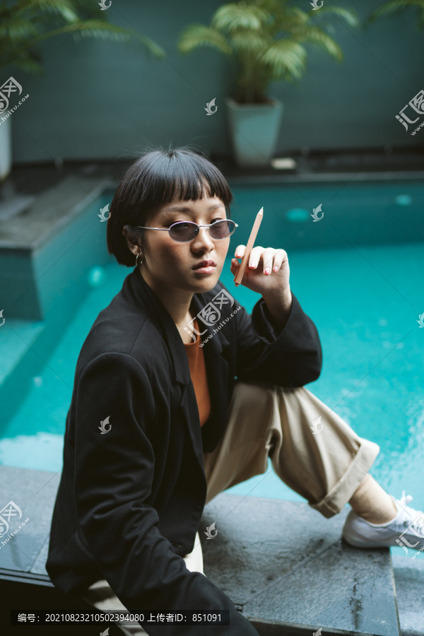 游泳池附近戴太阳镜的短发亚洲时髦女性肖像。她像香烟一样拿着钢笔。
