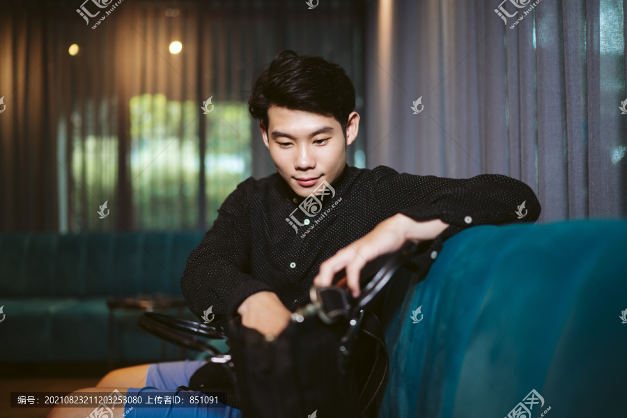 一个穿着黑衬衫的亚洲人坐在沙发上，在包里找东西。