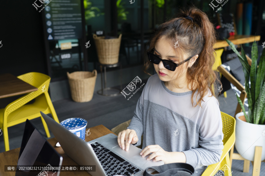美丽的亚洲女人戴着太阳镜在户外咖啡馆的笔记本电脑上工作。