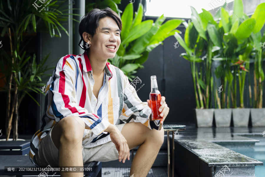 兴高采烈的亚洲年轻人坐在家里的游泳池旁，享受着一瓶美酒。