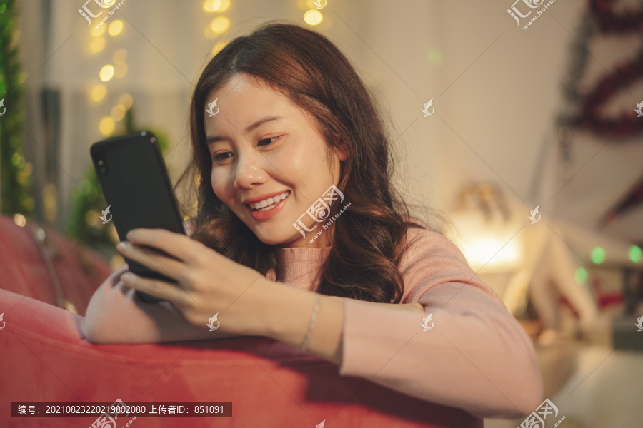 在圣诞节，年轻的亚洲美女在沙发上用智能手机梳理黑发。