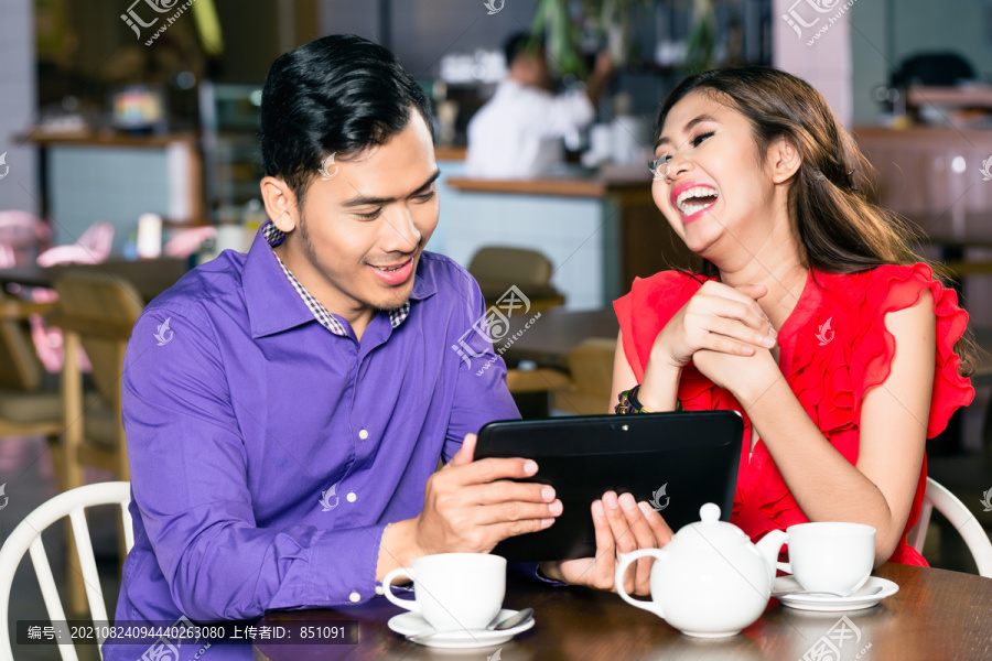在一家现代化的咖啡馆里，一位有幽默感的年轻帅哥在他的漂亮女友旁边的平板电脑上观看一段有趣的视频