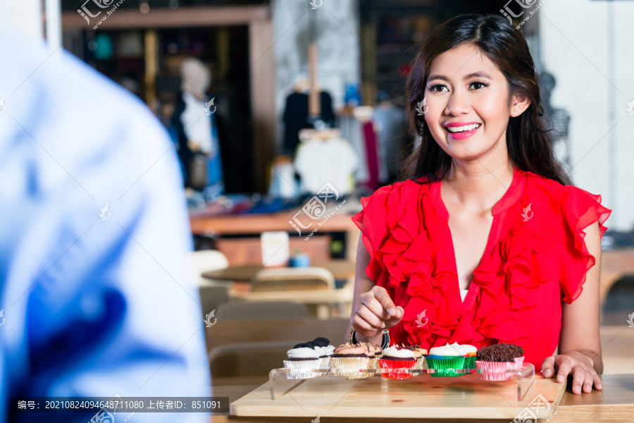 一位快活的亚洲妇女在凉爽的咖啡店柜台后点着纸杯蛋糕，里面有各种美味的甜点